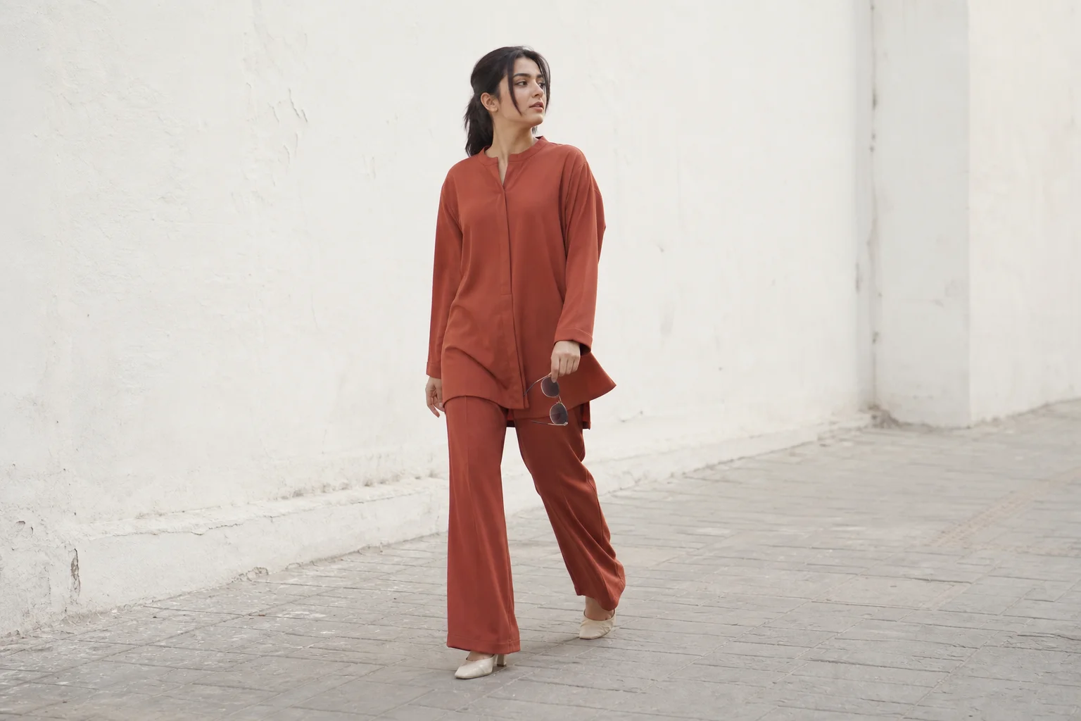 Young woman in a modern rust co-ord set walking past a minimal white wall, contemporary Pakistani street style